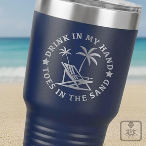 May include: Dark blue insulated travel mug with a silver lid. The mug has an etched design with the words "Drink in my hand, toes in the sand" around a beach scene. Ideal for cold drinks, it's a stylish travel accessory.