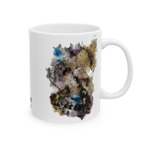 Könnte beinhalten: Weiße Keramiktasse mit einem farbenfrohen abstrakten Design in Blau-, Braun- und Goldtönen. Die Tasse hat einen gebogenen Henkel und eine glatte, glänzende Oberfläche. Das Design scheint im Aquarell- oder Tuschewaschstil gehalten zu sein.
