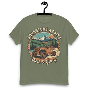 May include: Khaki green t-shirt with a graphic of an off-road vehicle, the text "Adventure Awaits" and "Dust & Glory". The design includes a mountain landscape with a setting sun and trees.