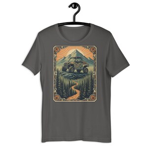 Könnte beinhalten: Dunkelgraues T-Shirt mit einer Grafik eines Geländewagens auf einem Bergpfad, mit einem Berg und der Sonne im Hintergrund. Das Design ist mit einem verzierten Rahmen versehen. Das T-Shirt hängt an einem schwarzen Kleiderbügel.