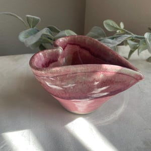 May include: A handmade, heart-shaped ceramic bowl with a glossy, mottled pink glaze. The bowl has a unique, organic form and a smooth interior. The exterior features variations in color, adding to its handcrafted appeal. Ideal for serving or as a decorative piece.