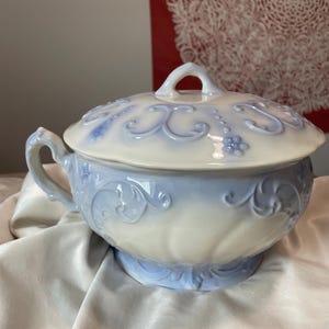 May include: A white and light blue ceramic tureen with a lid. The tureen has a handle on each side and a loop handle on the lid. The design features embossed floral and scroll patterns. The tureen sits on a pedestal base.
