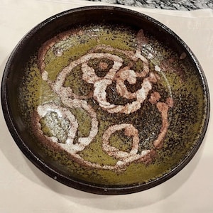 May include: A handmade ceramic bowl with a dark brown rim and a green interior. The bowl features a white and brown spiral design in the center. The bowl is round and has a textured surface.