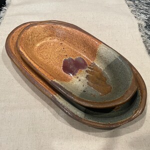 May include: Two nested oval ceramic dishes with a speckled finish. The interior features a warm orange-brown tone with a dark maroon accent and a contrasting grey-green section. The outer edges are a deep brown, showcasing a handcrafted texture.