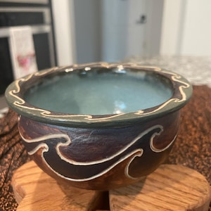 May include: A handcrafted ceramic bowl with a dark brown exterior and a light blue interior. The bowl features a wave design in white and is set on a wooden trivet. The rim has a green border with a white wave pattern.