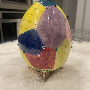 May include: A decorative egg-shaped object with a patchwork design in yellow, purple, blue, and red. The egg is adorned with rhinestones and rests on a small, decorative stand. The egg is a unique home decor item.
