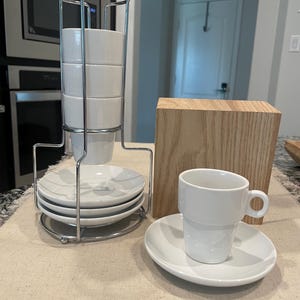 May include: A white ceramic espresso cup and saucer set. The cup has a handle and is stacked on a matching saucer. A set of four stacked cups and saucers are held in a chrome metal rack. A wooden block is in the background.