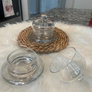 May include: A clear glass cloche with a knob on top, sitting on a woven, circular placemat. Beside it are two additional glass pieces, one a base and the other a tilted bowl-shaped piece. The items are on a white, fluffy surface.