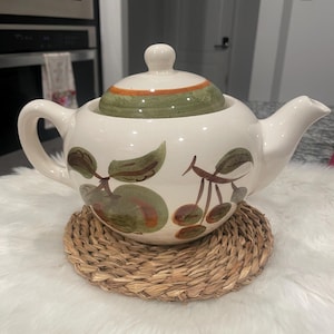 May include: A white ceramic teapot with a lid, handle, and spout. The teapot is decorated with green apples and brown cherries. It sits on a round, woven, natural-colored placemat.