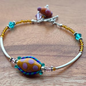 Puede incluir: Pulsera hecha a mano con una cuenta central amarilla con lunares morados, enmarcada por detalles azules y verdes. Incluye cuentas naranjas, amarillas y turquesas, y un cierre plateado. La pulsera está ensartada en un cordón blanco.