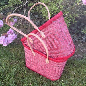 Peut inclure: Deux paniers roses et rouges tressés avec des anses beiges sont posés sur de l'herbe verte. Les paniers ont une forme rectangulaire avec un motif tissé. Les bords sont d'une nuance de rouge plus foncée. Les anses sont en beige clair.