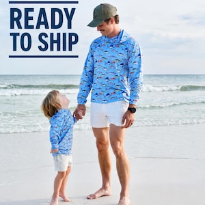 Könnte beinhalten: Zwei langärmelige Kapuzenpullover mit hellblauem Hintergrund und einem sich wiederholenden Muster aus bunten Fischen, kombiniert mit weißen Shorts. Der Text READY TO SHIP ist dunkelblau auf der linken Seite abgebildet.