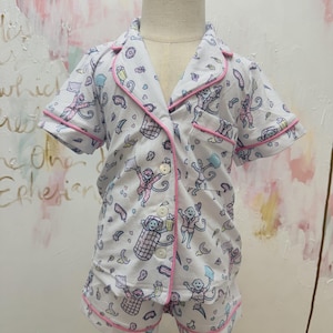 May include: A white short-sleeve pajama set with a button-down top and matching shorts. The fabric features a repeating pattern of monkeys, pillows, and stars in blue and pink, accented with pink piping along the collar, pocket, and cuffs.