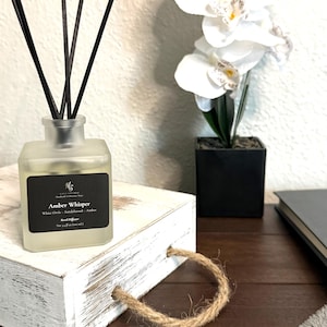 Puede incluir: Un frasco difusor de varillas de vidrio esmerilado con varillas negras y una etiqueta negra que dice Amber Whisper, White Orris, Sandalwood, Amber y Net 3.4 fl oz (100 ml). El frasco se asienta sobre una caja de madera blanca desgastada con un asa de yute.