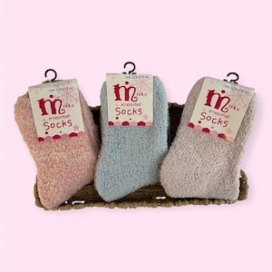 May include: Three pairs of fuzzy socks in pink, blue, and beige, displayed in a woven basket. Each pair has a tag that reads "minky accessories Socks" and "ONE SIZE FITS ALL". The socks are soft and plush.