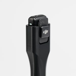 Peut inclure: Un microphone sans fil noir avec une pince et le logo DJI. Le microphone a une petite forme rectangulaire avec un dessus arrondi et une série d'ouvertures. Une petite lumière verte est visible sur le dessus du microphone.