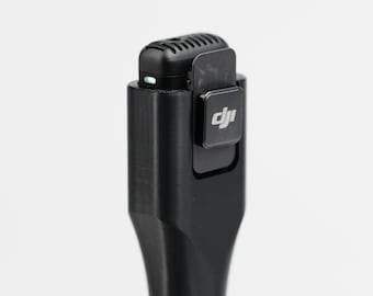 May include: A black wireless microphone with a clip and the DJI logo. The microphone has a small, rectangular shape with a rounded top and a series of vents. A small green light is visible on the top of the microphone.