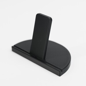 May include: A black, semi-circular display stand with a vertical support. The stand is made of a solid, matte material. The vertical support is rectangular and positioned in the centre of the base. The base is approximately 10 cm wide.
