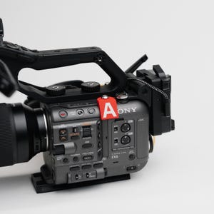 May include: A professional-grade digital cinema camera with a large lens and a black handle. The camera body is gray with various buttons and dials. A red tag with the letter "A" is attached to the camera.