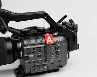 May include: A professional-grade digital cinema camera with a large lens and a black handle. The camera body is gray with various buttons and dials. A red tag with the letter "A" is attached to the camera.