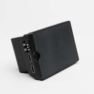 May include: A black electronic device with a textured surface and multiple ports. The device has a rectangular shape with a slightly angled side, featuring two HDMI ports and other connection points.