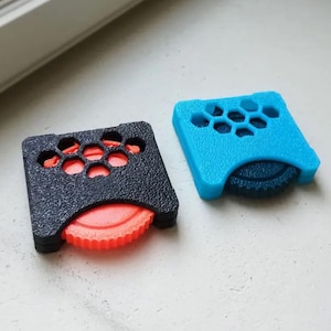 Peut inclure: Deux petits objets carrés imprimés en 3D. L'un est noir avec des accents orange, l'autre est bleu avec des accents bleu foncé. Les deux ont un motif en nid d'abeille sur le dessus et un composant circulaire et strié visible sur le côté. Ce sont des petits outils ou accessoires.