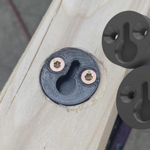 May include: Three black circular keyhole hangers made of plastic. One hanger is installed into a light-colored wooden board using two gold-colored star-drive screws. Two additional identical hangers are shown floating to the right of the board.