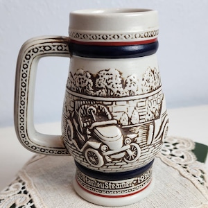 May include: A vintage ceramic beer stein with a detailed relief design featuring a classic car and the text "Stanley Steamer 1936". The stein has a handle and is decorated with blue and red bands. It rests on a green book with a lace doily.
