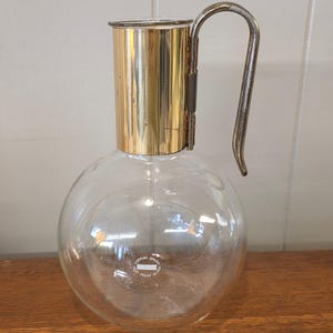 May include: A clear glass carafe with a round body and a gold-colored metal top and handle. The handle is a curved, silver-colored metal. The carafe has a label that reads "Corning Brand Heat Proof Glass".