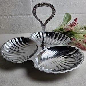 May include: A silver-toned metal serving dish featuring three distinct shell-shaped compartments arranged in a clover pattern. A central vertical handle with an ornate, textured loop design rises from the middle, connecting the three sections.