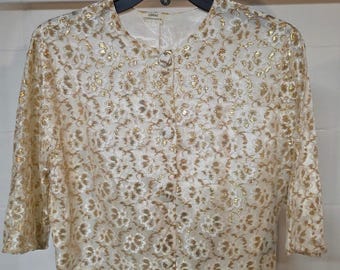 May include: A cream-colored cropped jacket featuring a metallic gold floral lace overlay. The garment has three-quarter length sleeves, a round neckline, and a front button closure with three matching fabric-covered buttons.