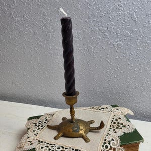 May include: A dark purple twisted taper candle held in a brass candle holder shaped like a turtle. The turtle base features a detailed shell pattern and four legs, with the candle holder stem rising from the center of the turtle's back.