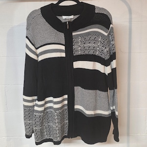 May include: A black, white, and gray zip-up cardigan. The sweater features a variety of patterns, including stripes and geometric designs. The long-sleeved cardigan is hanging on a black hanger against a white brick wall.
