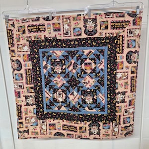 May include: A square quilt featuring a central panel with blue, black, and peach geometric patterns and illustrations of figures in chairs. The border displays a repeating peach background with cherries, bowls, and the text Life is just a chair of bowlies.