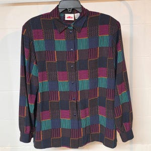 May include: A long-sleeve button-down shirt featuring a multi-colored checkered pattern in shades of black, magenta, teal, and blue. The shirt has a pointed collar, a front button placket with dark buttons, and a label at the neck reading Alia.