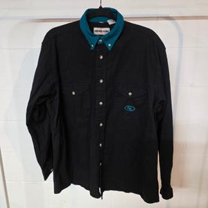 May include: Black long-sleeve button-down shirt with a teal collar and cuffs. The shirt has two chest pockets and a teal embroidered logo on the front. The collar has the text "COWBOY CODE".