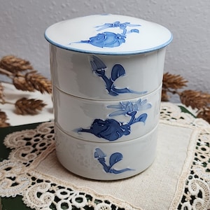Puede incluir: Un recipiente de cerámica blanco de tres niveles con tapa. El recipiente está decorado con diseños florales azules. La tapa y los bordes tienen un ribete azul. El recipiente está sobre un tapete de encaje color crema.