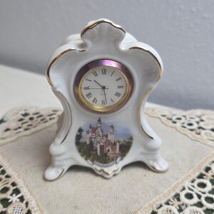 May include: A white ceramic mantel clock with gold trim. The clock face has Roman numerals and gold hands. A colorful image of a castle is below the clock face. The clock is on a beige surface.