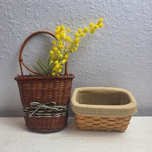May include: Two woven baskets. One is a dark brown basket with a handle and a green and white striped ribbon. The basket contains yellow flowers. The other basket is light brown with a beige fabric lining.