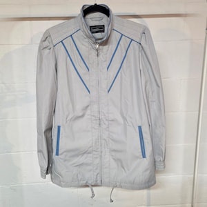 May include: A light gray jacket with blue accent stripes and a matching collar. The jacket has a zipper closure, two front pockets, and a drawstring at the bottom. The jacket is hanging on a black hanger against a white wall.