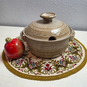 May include: A ceramic soup tureen with a lid, featuring a floral design, sits on a floral placemat. A red, apple-shaped decorative item is to the left. The tureen is beige with a dark opening for a ladle.