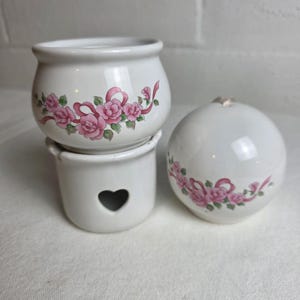 May include: A white ceramic oil warmer set featuring a bowl and a base with a heart-shaped cutout. Both pieces are decorated with a pattern of pink roses and green leaves intertwined with pink ribbons. A matching white ceramic sphere is included.