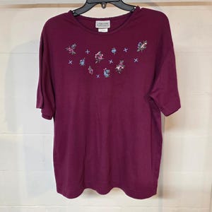 May include: A short-sleeved, burgundy-colored shirt with embroidered floral designs and small blue crosses. The shirt has a round neckline with a scalloped edge and a tag that reads "Janzen The Classics."