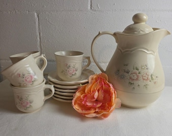 May include: A cream-colored ceramic tea set featuring a large pitcher with a lid and matching cups and saucers. Each piece is decorated with a delicate floral pattern of soft pink roses and green leaves, accented by a scalloped edge design.