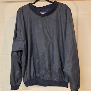 May include: A navy blue pullover windbreaker with a small checkered pattern. The windbreaker has a navy blue collar, cuffs, and waistband. The garment is hanging on a black hanger against a neutral background.