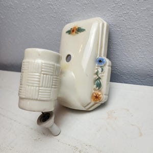 May include: Off-white ceramic bathroom set with floral accents. The set includes a rectangular soap dish and a cylindrical toothbrush holder with a woven pattern. The soap dish has a hole and is decorated with blue, orange, and green flowers.