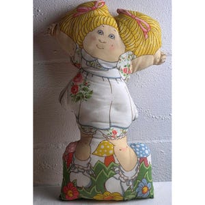May include: A soft fabric doll featuring a printed design of a child with blonde hair in pigtails, wearing a white dress with floral accents. The doll is depicted standing in a field of colorful mushrooms and flowers with a green grassy base.