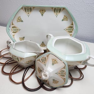 May include: A vintage porcelain tea set with a tray, creamer, sugar bowl, and lid. The set features a white base with a light green trim and gold and green floral designs. The tray measures approximately 12 inches long.