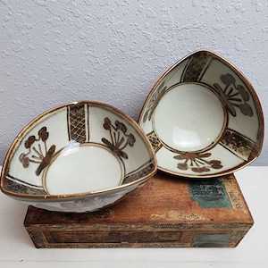 May include: Two triangular ceramic bowls with a white interior and a brown floral design. The bowls have a brown rim and are placed on a wooden box. The bowls are a unique shape and design.