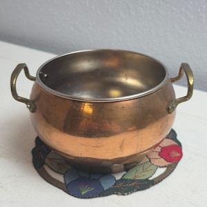 May include: A small copper pot with brass handles, resting on a floral-patterned coaster. The pot has a shiny interior and a slightly aged exterior. The coaster features a design of colorful flowers and leaves.